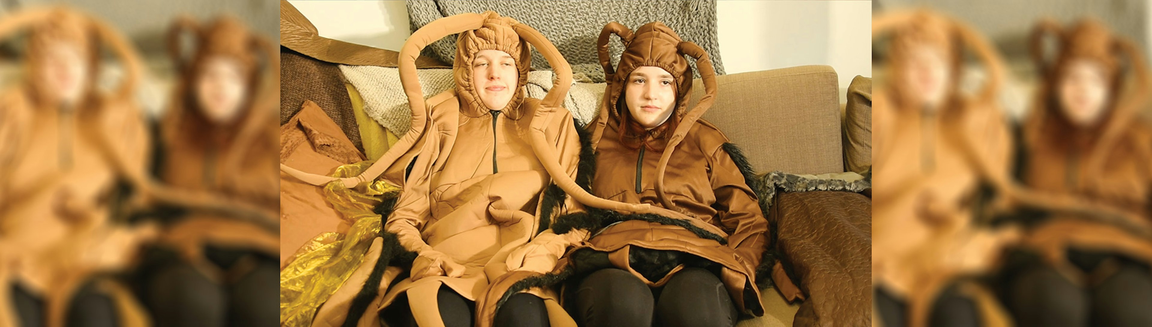 Two kids are sitting side by side on a couch dressed up in cockroach costumes