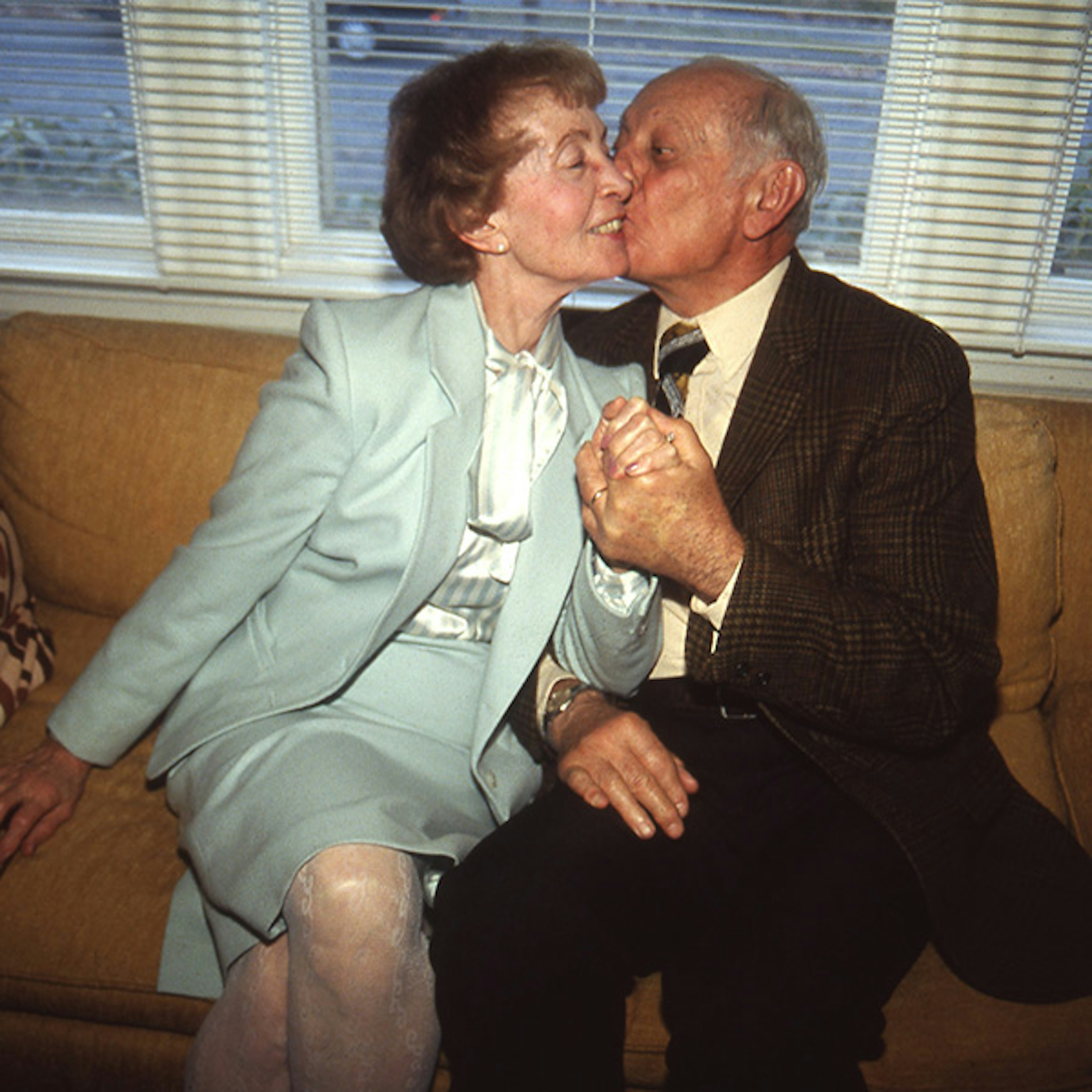 An elderly couple sitting on a couch at their 50th wedding anniversary, the man is wearing a suit and is holding his wife's hand while kissing her on the cheek both are smiling happily