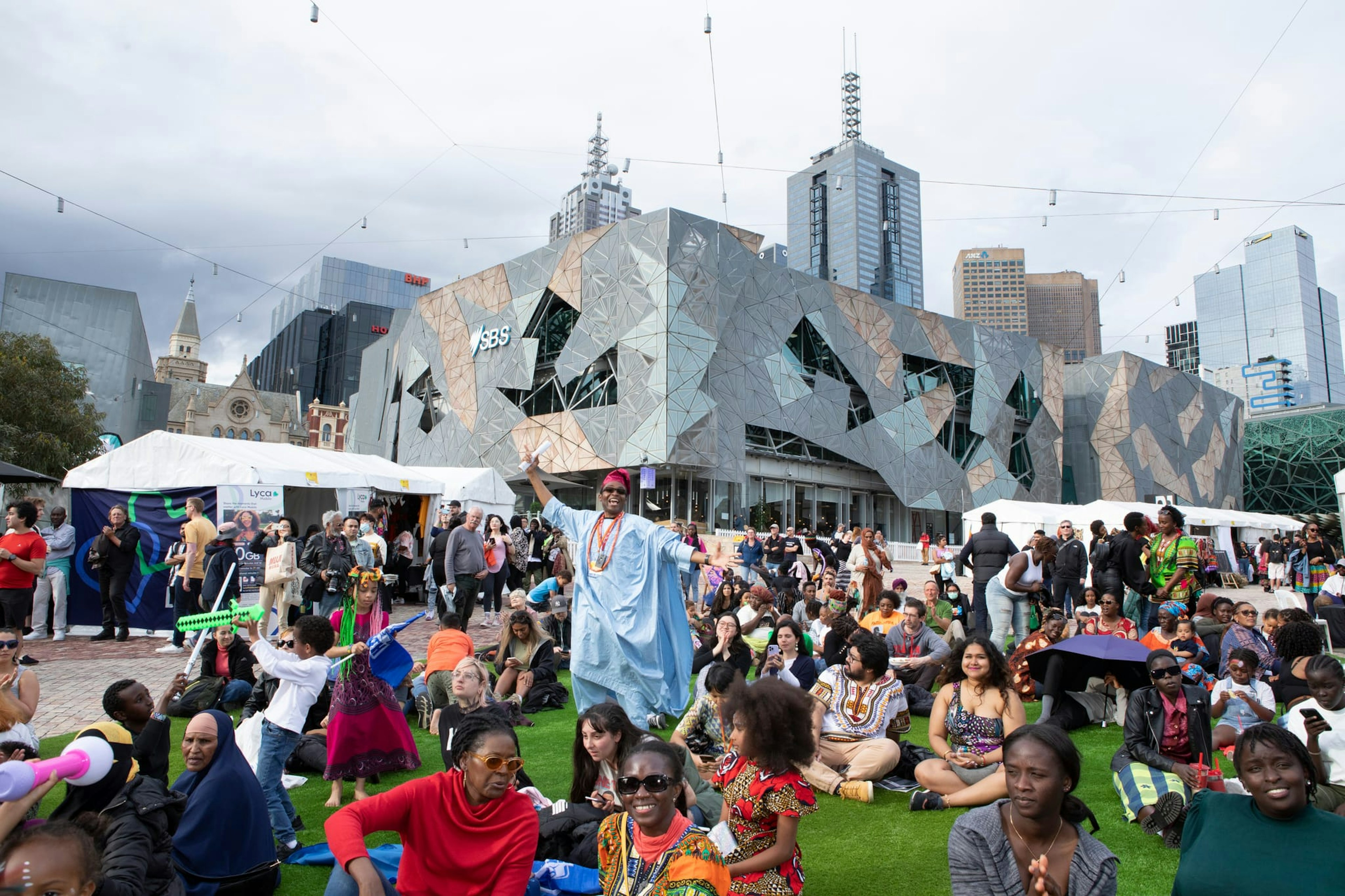 A crowd of people at Fed Square sit on grass, a man in the middle is dressed in a long blue robe.