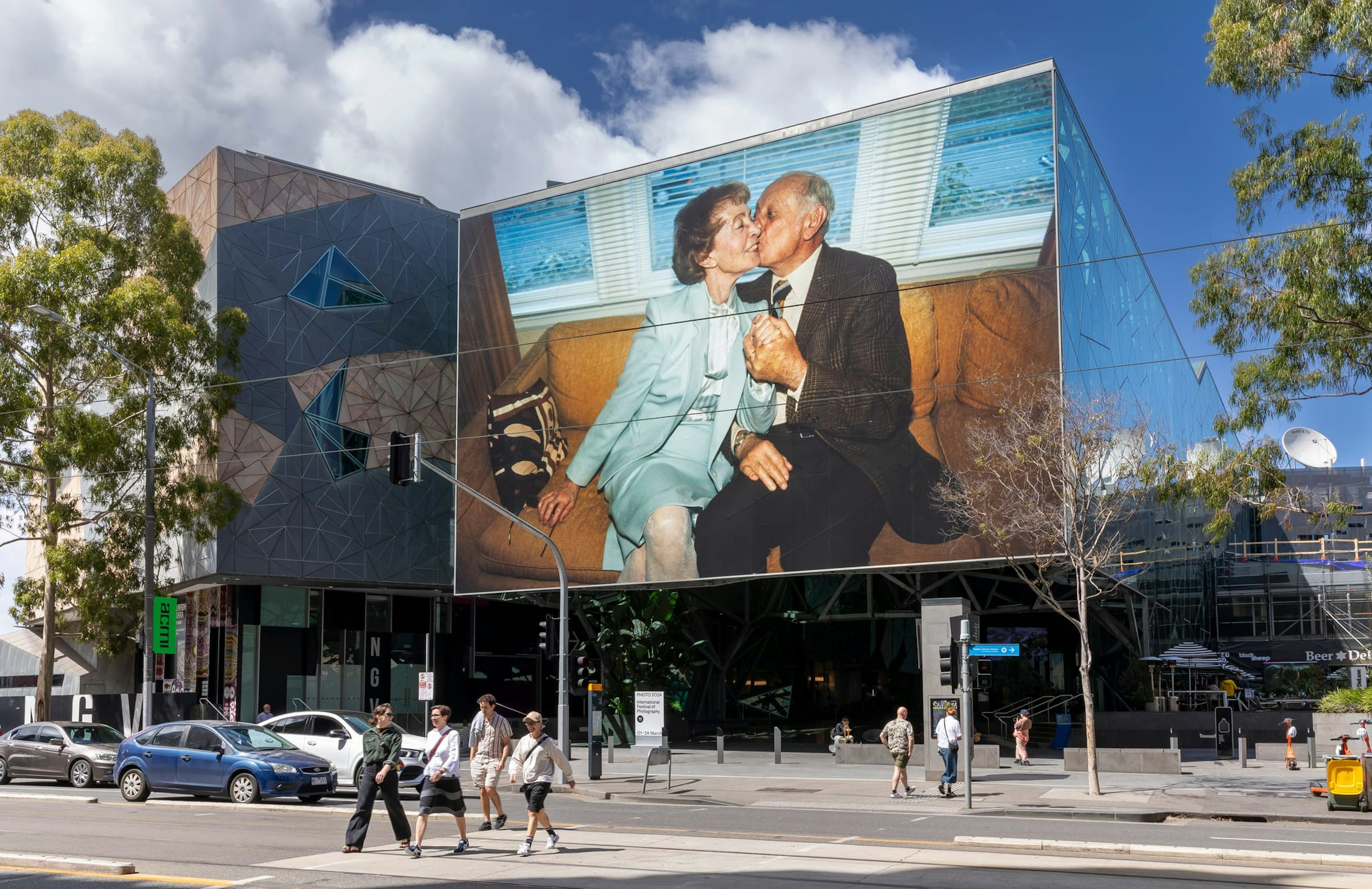 A large photo of two elderly people sitting on a couch, the man kissing the woman on the cheek, adorns the exterior of Fed Square's The Atrium. The photo is very large. People walk across the road at the lights, below the image. It's a sunny day with a blue sky and some white clouds.