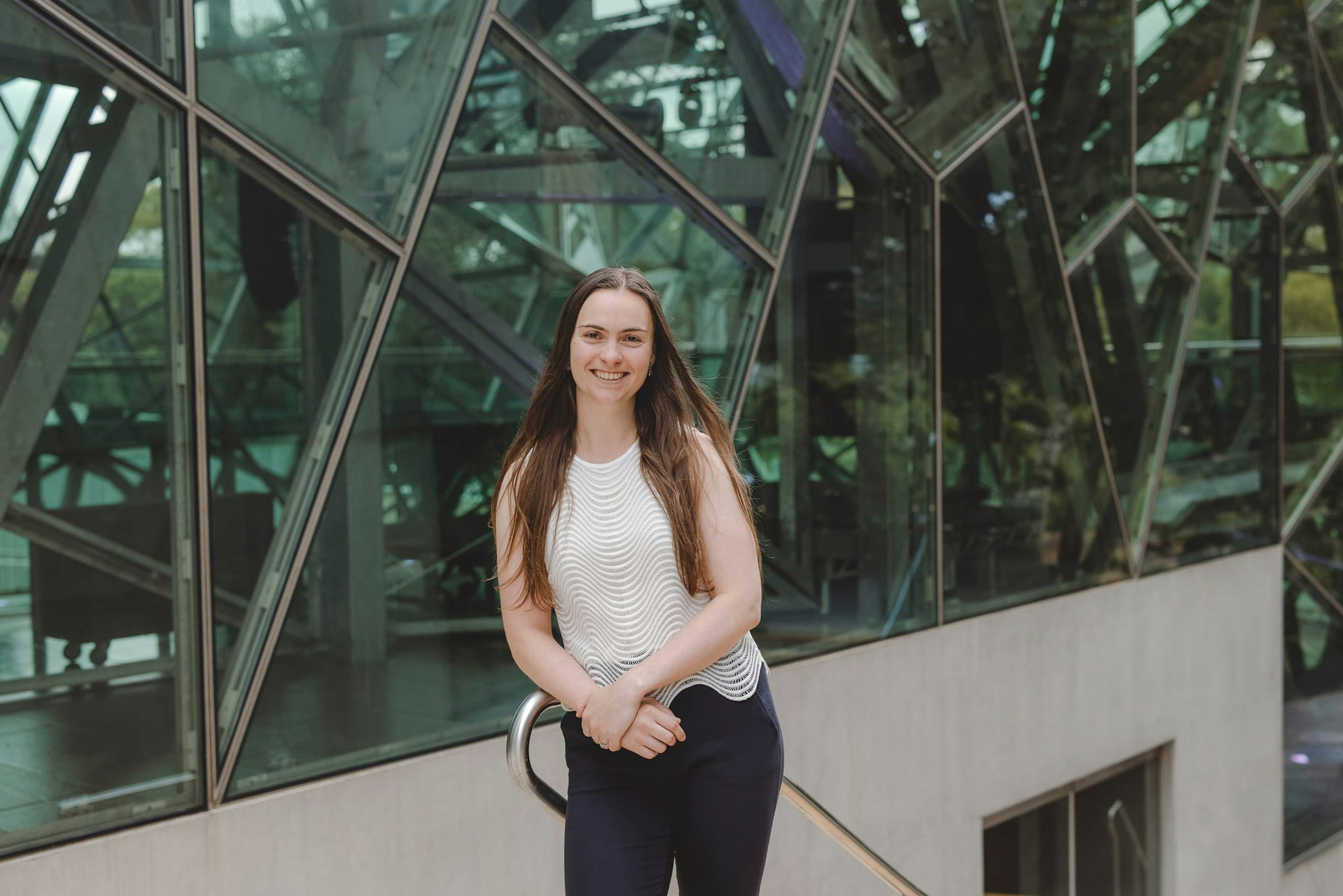 A woman with long brown hair looks at the camera, smiling. She wears a white top and black pants and her hands are crossed. She leans against a ballustrde and behind her are the glass fractals of the architecture at Fed Square.