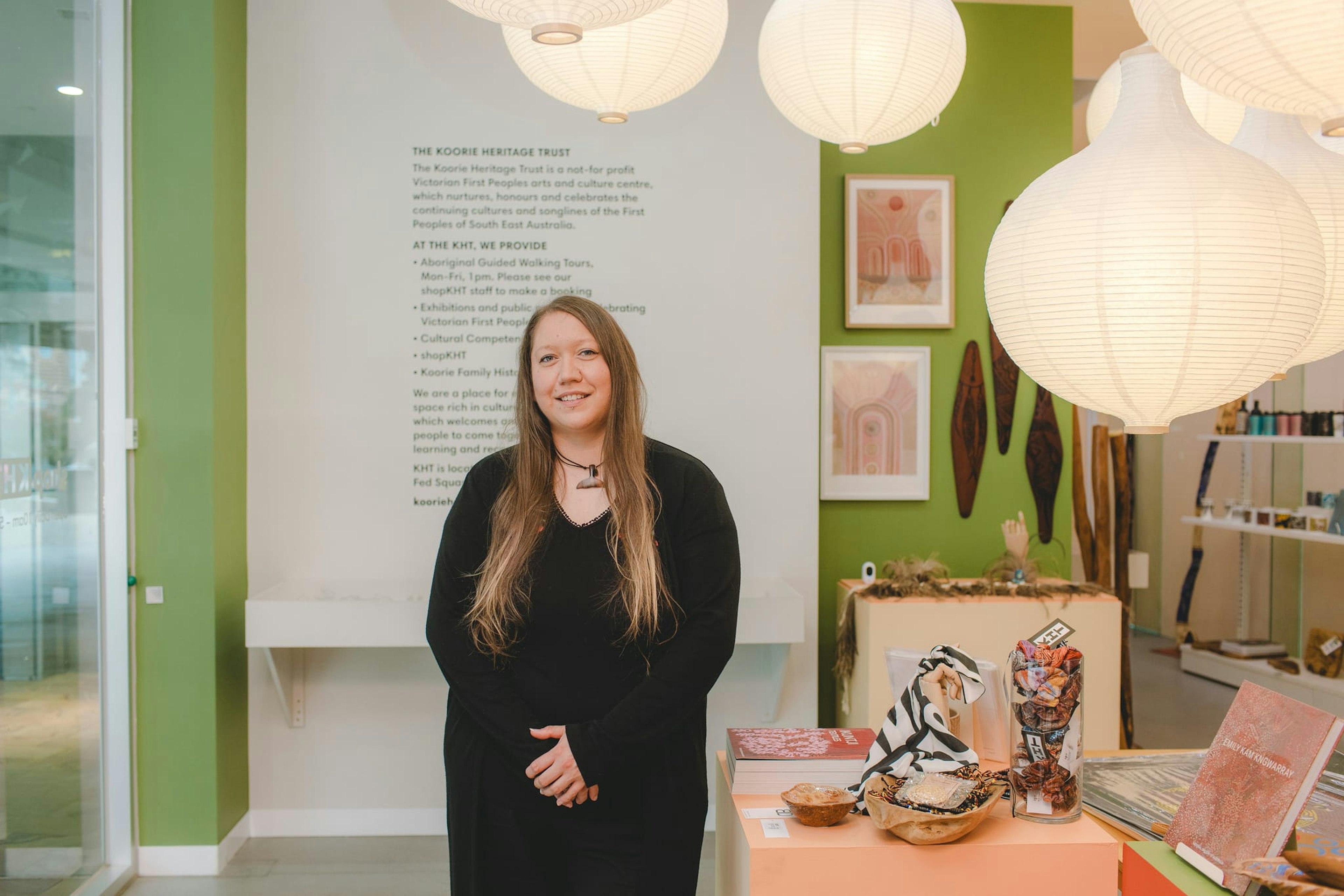 A woman with long dark-blonde hair smiles at the camera. She wears a long black cardigan, with her hands folded in front of her. She stands in a shop, near some colourful pieces, with large paper lanterns above her emitting a soft light. The wall behind her is green, with writing on it.