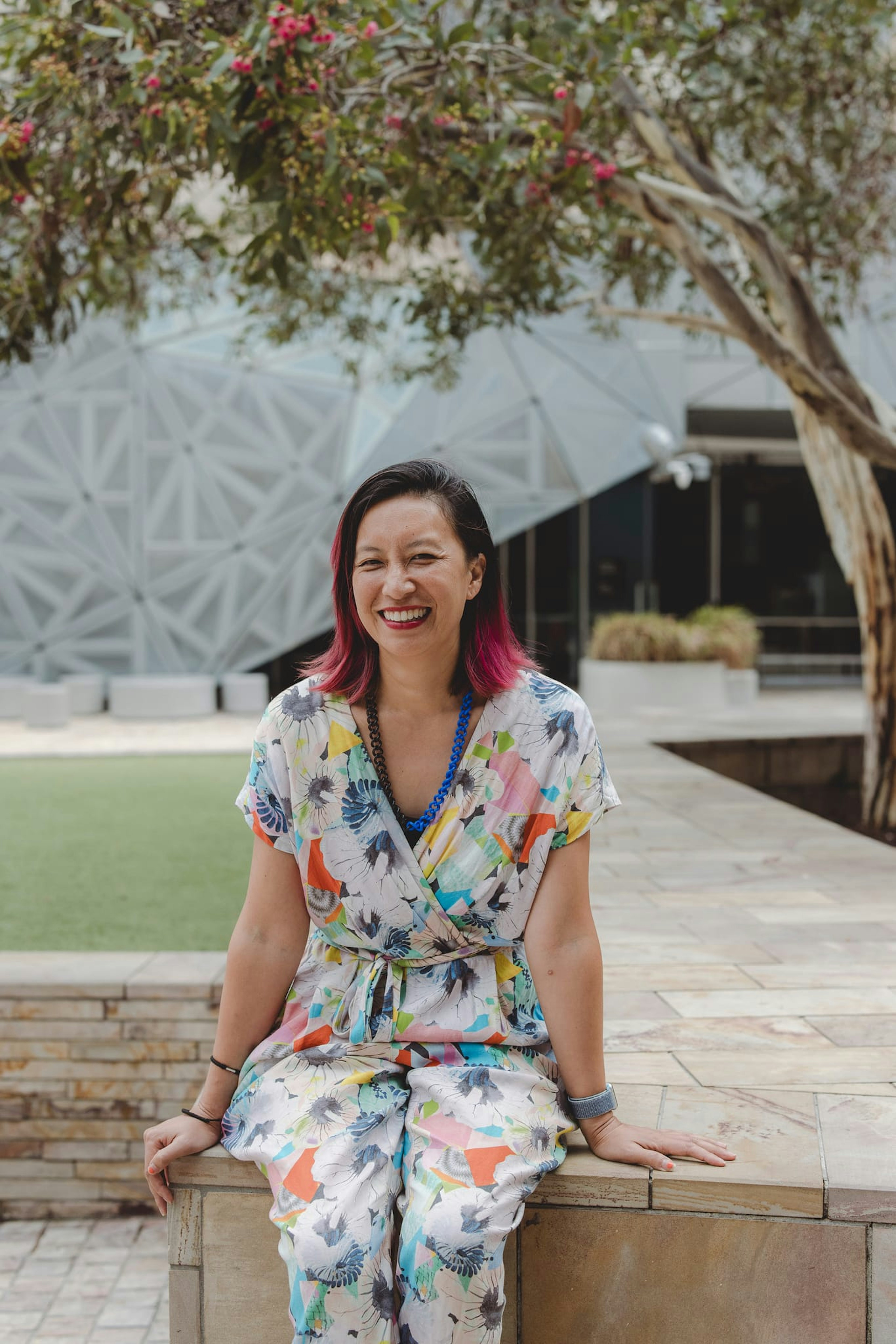 A woman with coloured pink hair smiles at the camera, her hands in the pockets of her brightly coloured jumpsuit. She sits on a coloured sandstone low wall, and green leaves of a gum tree are above her head. We see the grey fractal architecture of Fed Square behind her.