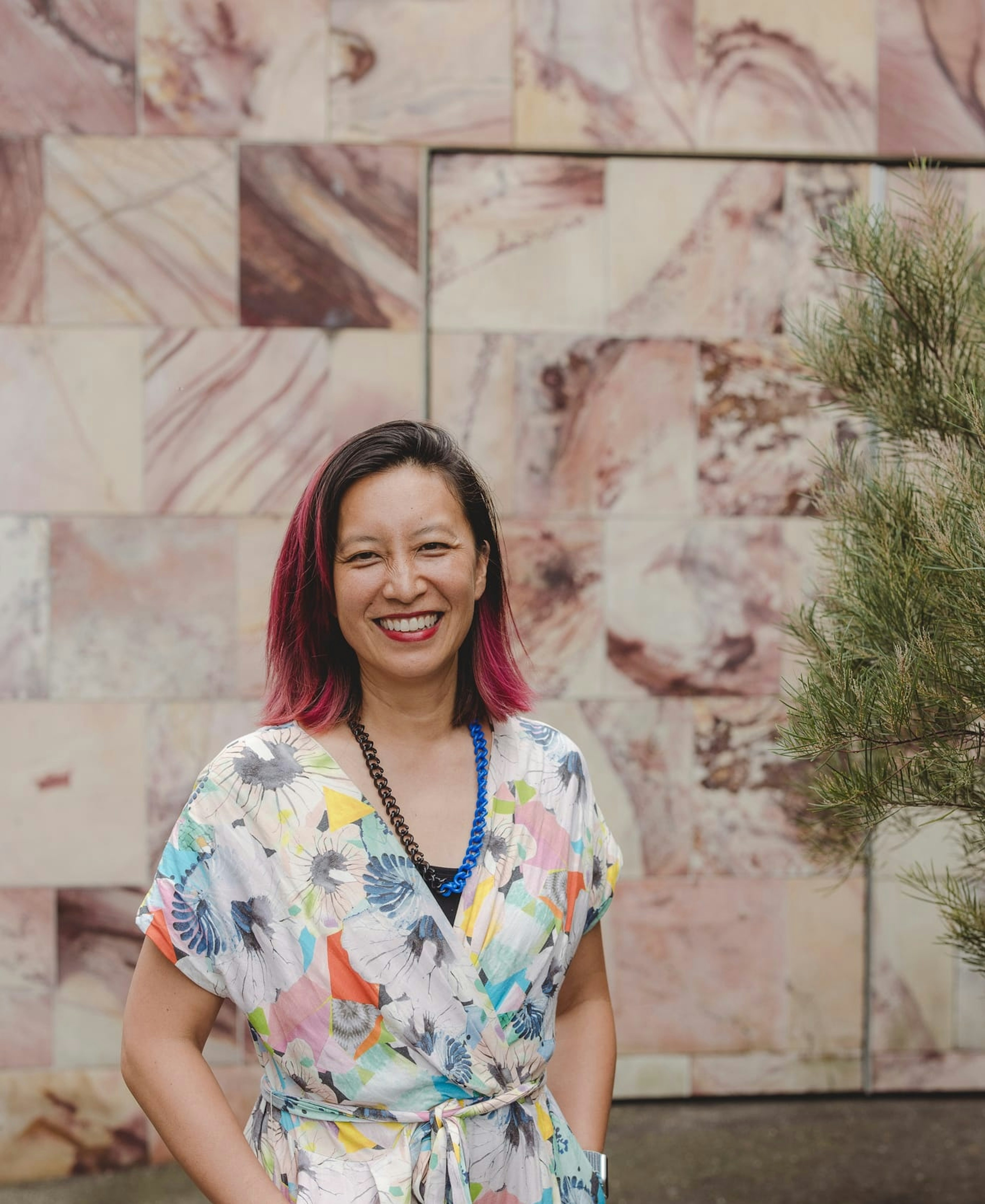 A woman with coloured pink hair smiles at the camera, her hands in the pockets of her brightly coloured jumpsuit. She stands against a coloured sandstone wall, and the soft green foliage of a plant is to her left.