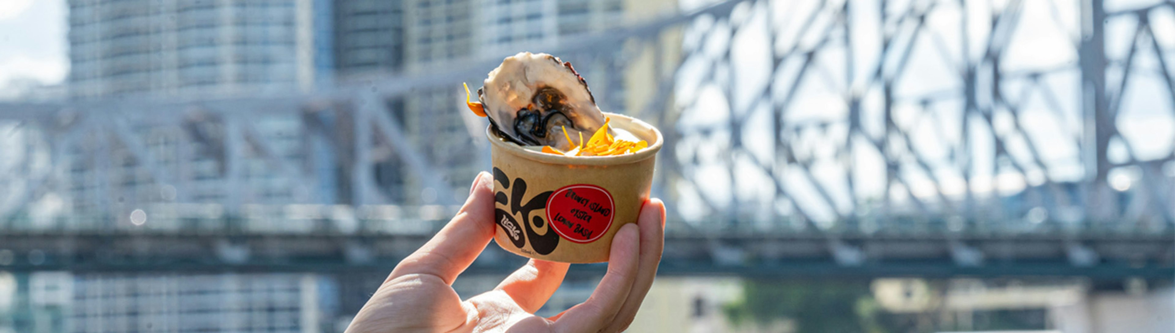 A hand holds a scoop of icecream in a cup, with an oyster in it. A bridge is in the background