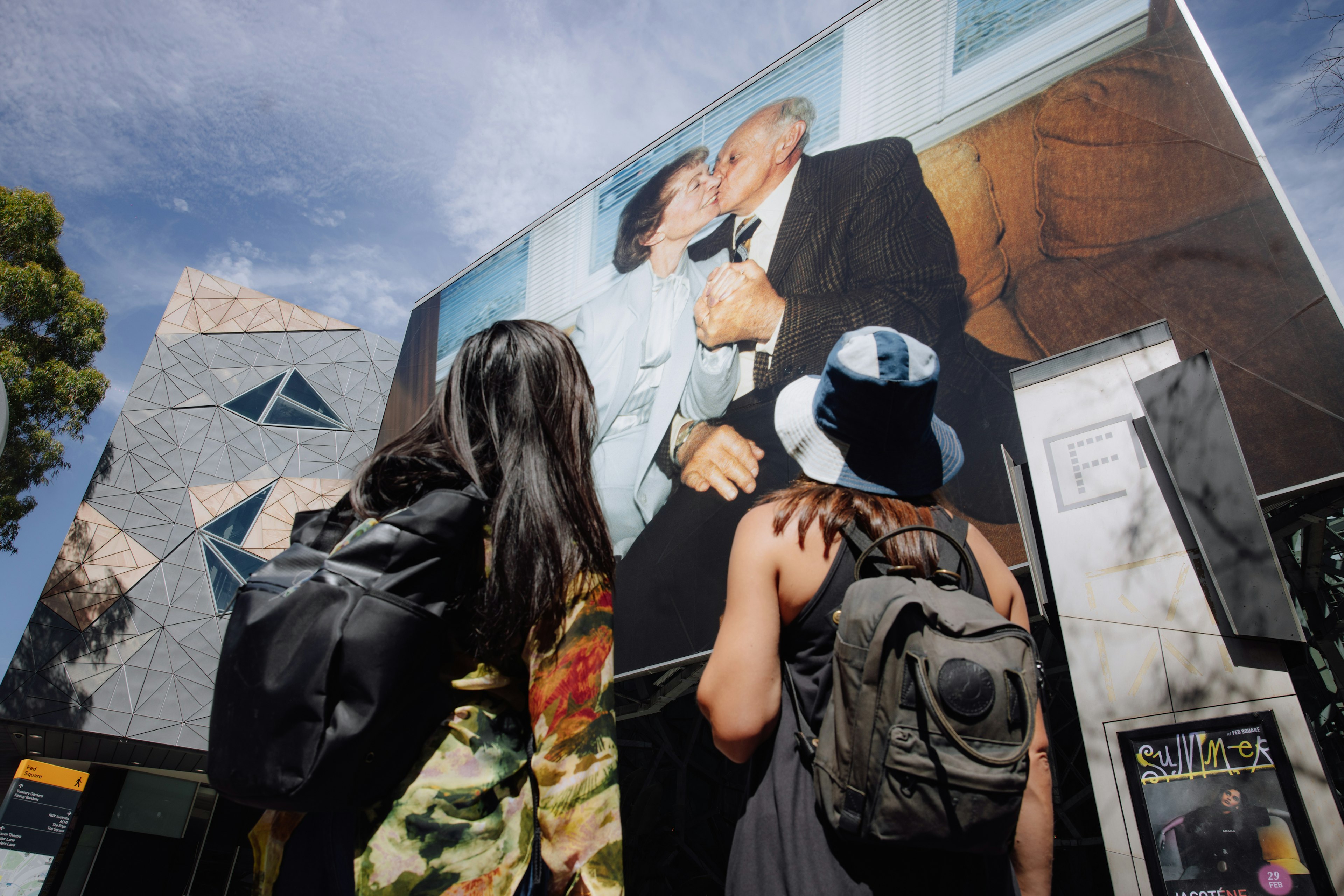 Two women look up to the enormous installation on the exterior of the Atirum, of two elderly people sitting on a couch. The sky is blue. The man in the image kisses the woman on the cheek.