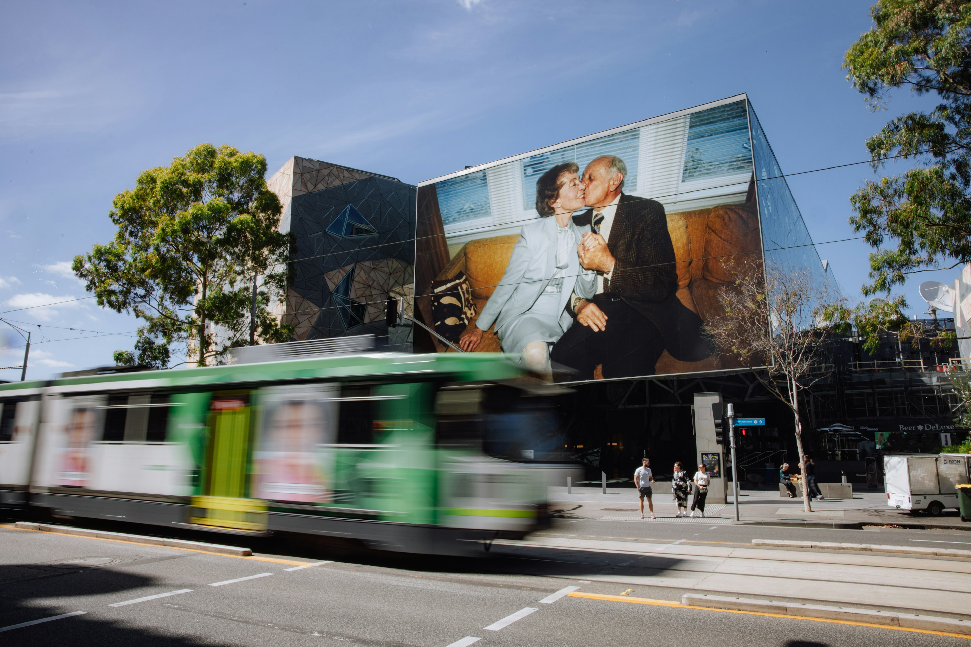 A tram rolls down Flinders Street, and on the exterior of the Atrium a large-scale installation of a photo is of an elderly couple sitting on a couch. The man in the photo kisses the woman on the cheek.