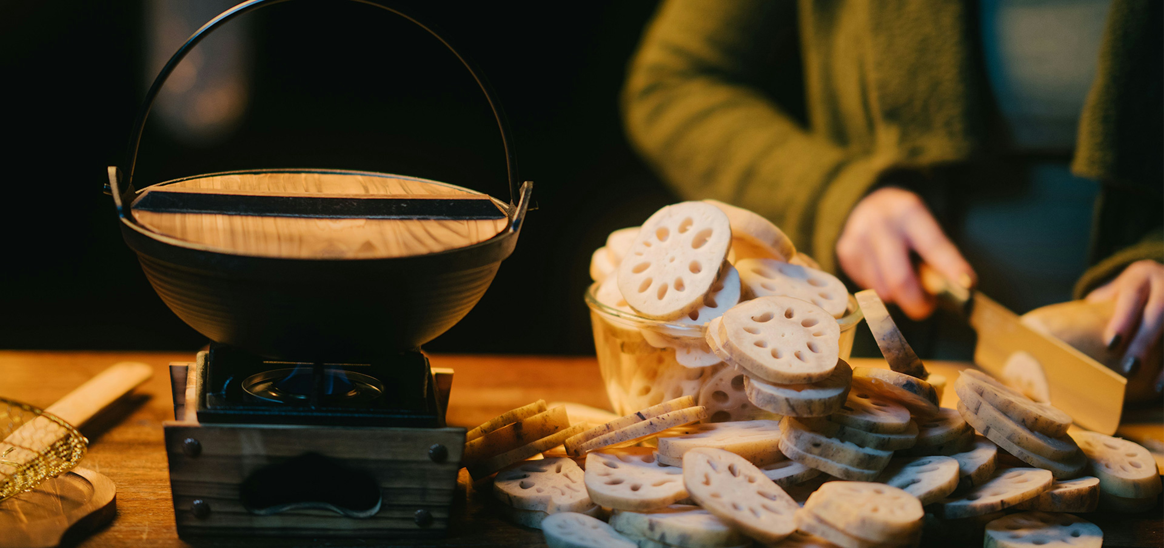 A photo of many lotus roots being cut up by a woman's torso that is out of focus with a pot on a small gas burner next to the lotus roots