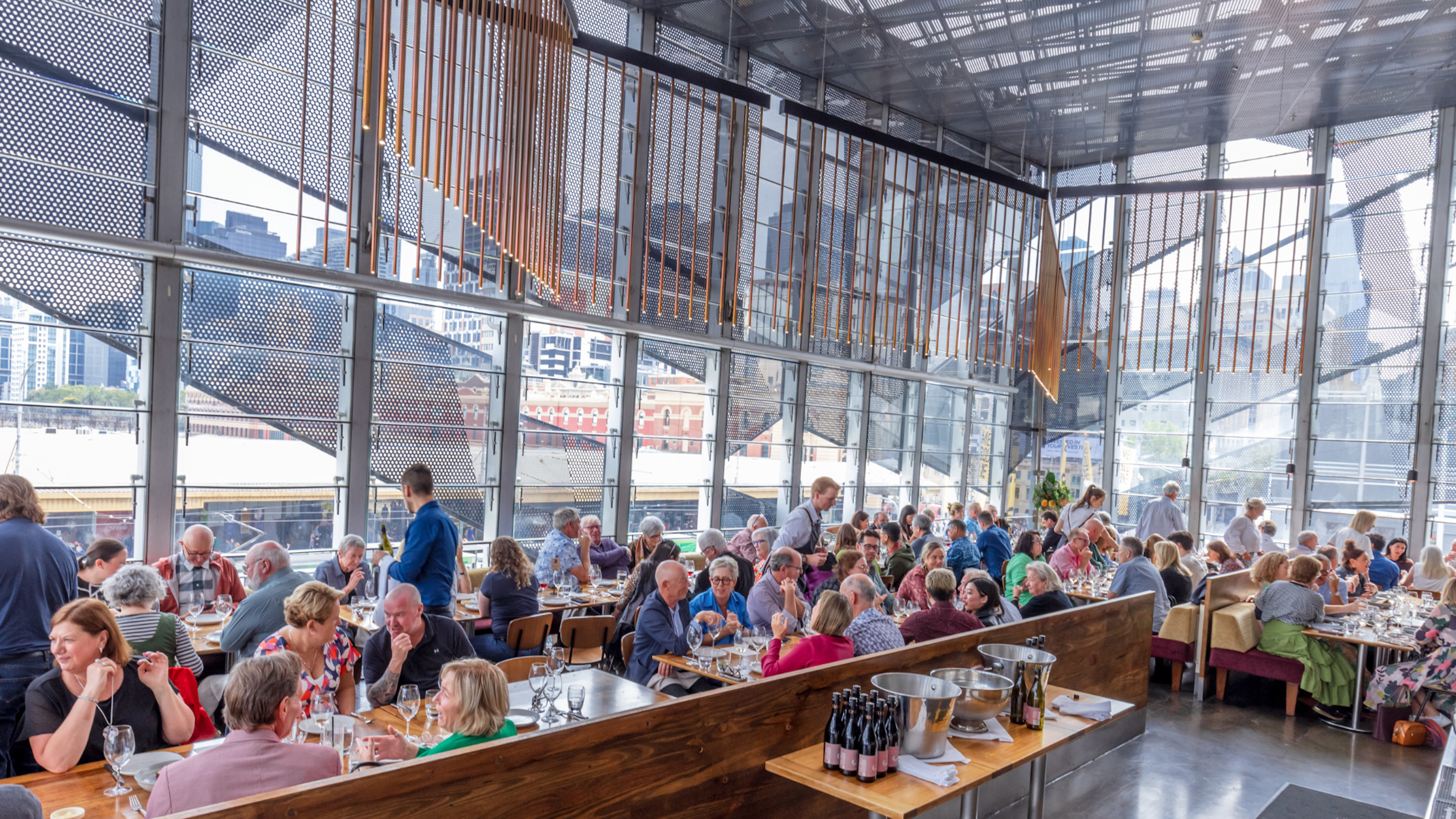 Many people sit at long timber tables at Taxi Kitchen, framed by floor to ceiling windows looking out to the city. It's a sunny day outside.