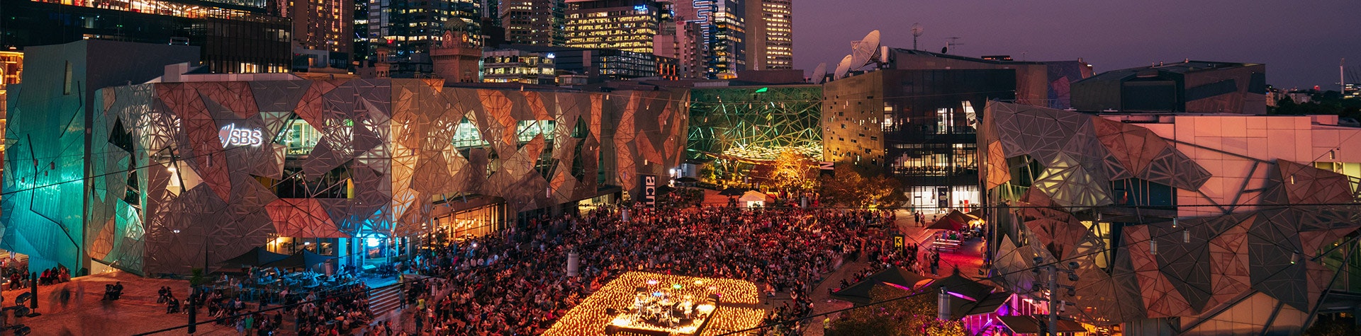 A crowd of people in the centre of Fed Square sitting around a stage that is surrounded by candles.