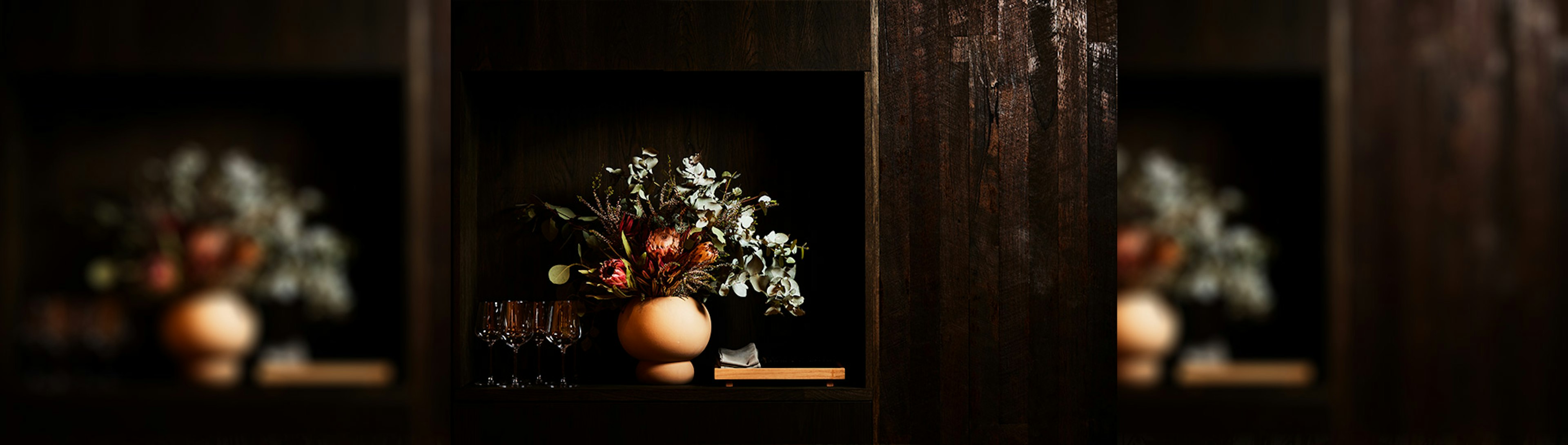 A vase sitting on a dark wood bench with Australian native flowers next to some wine glasses