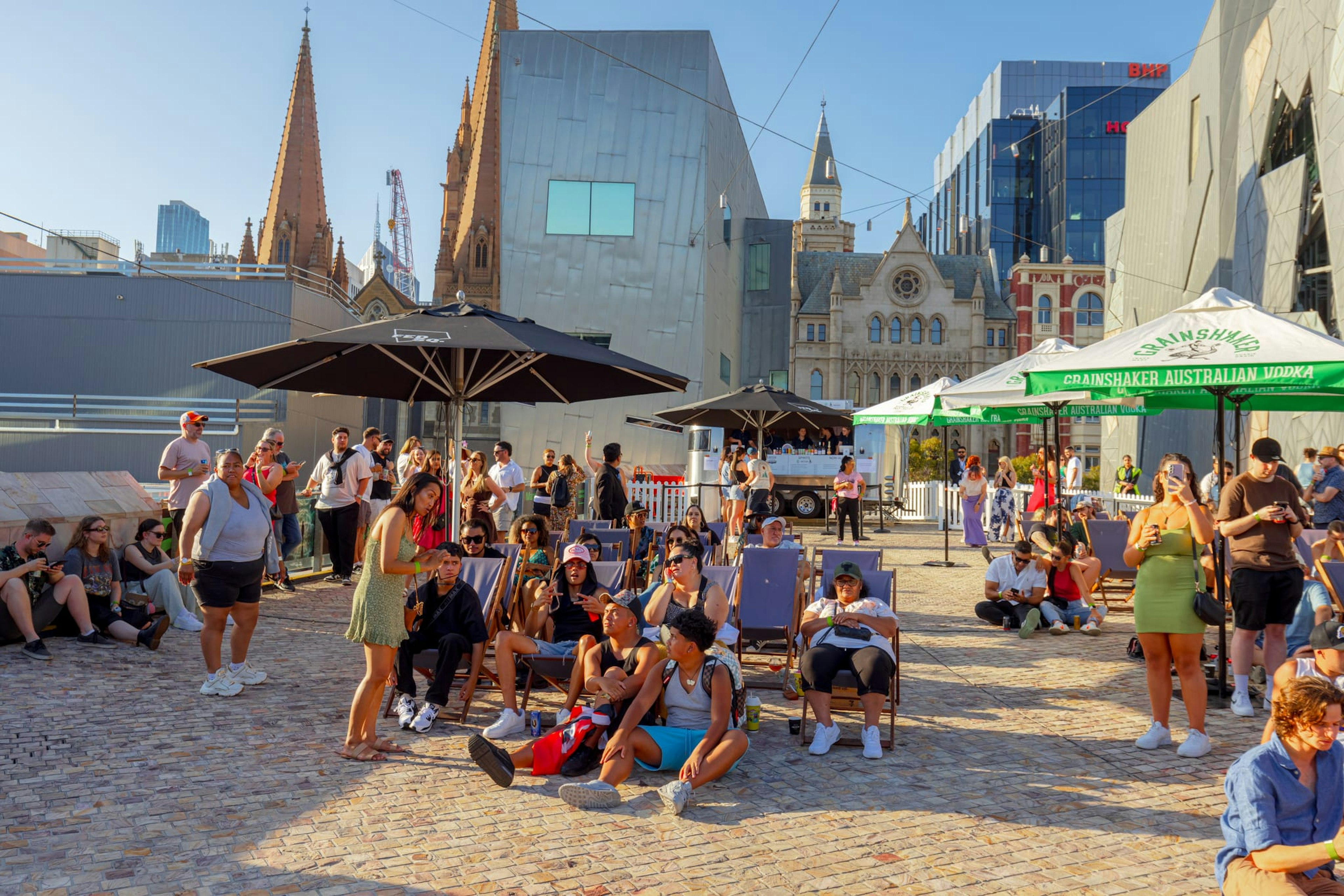 Umbrellas and deck chairs are set up in Fed Square outdoors at a pop-up bar, with a large group of attendees