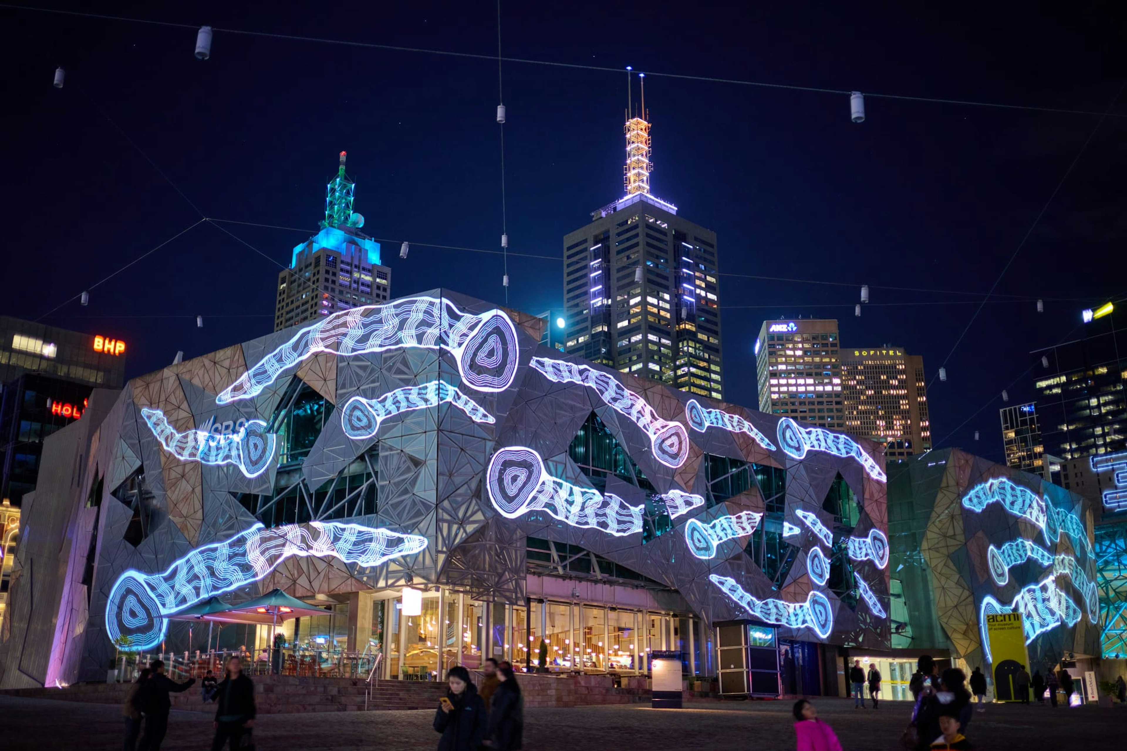 Images of white. glowing, line-drawn eel traps are projected onto Fed Square's buildings at night.