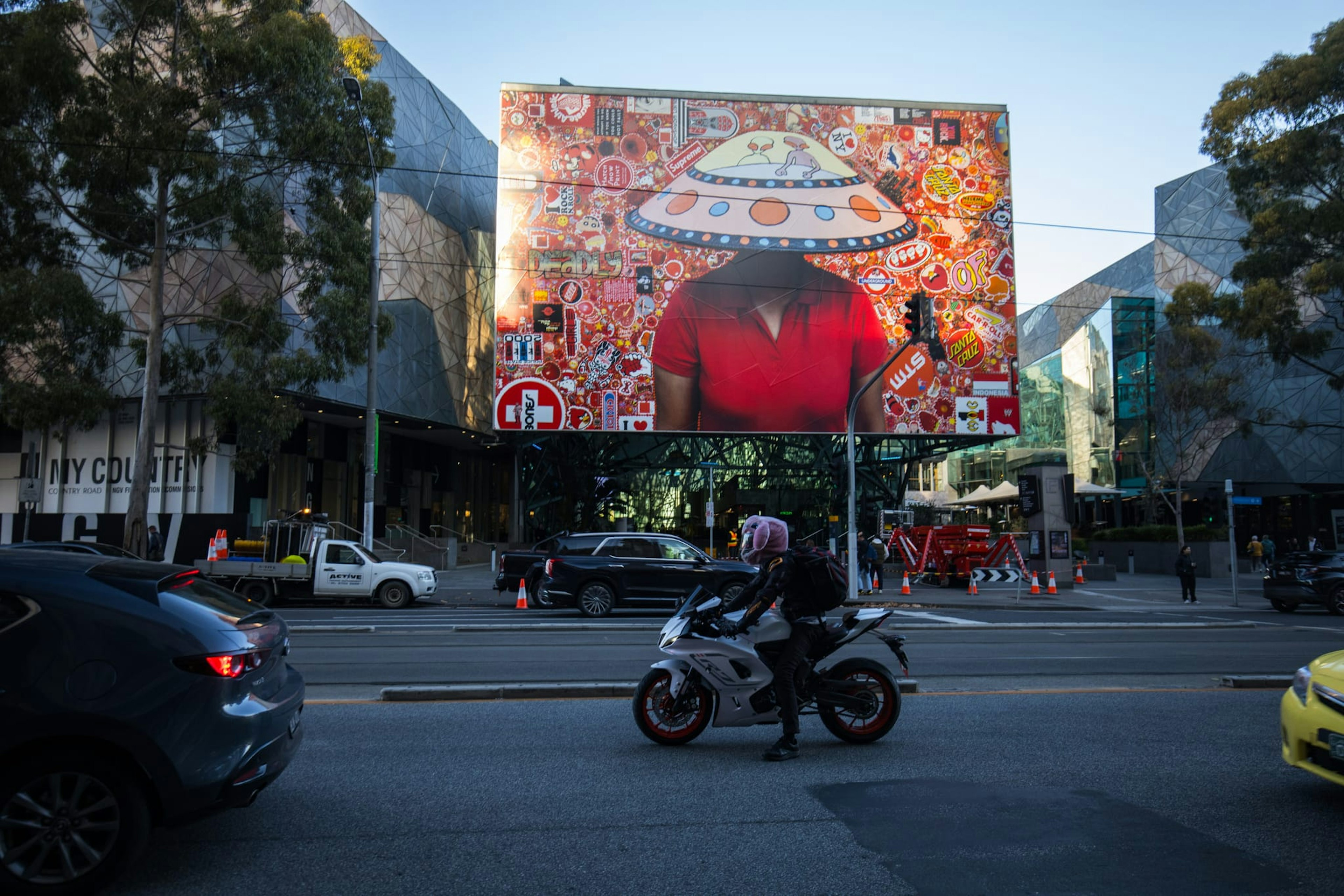 A large installation of a picture of a person wearing a UFO hat is decaled across the Atrium exterior at Fed Square. The view is from across the road, and a motorbike rides past in front of the image.