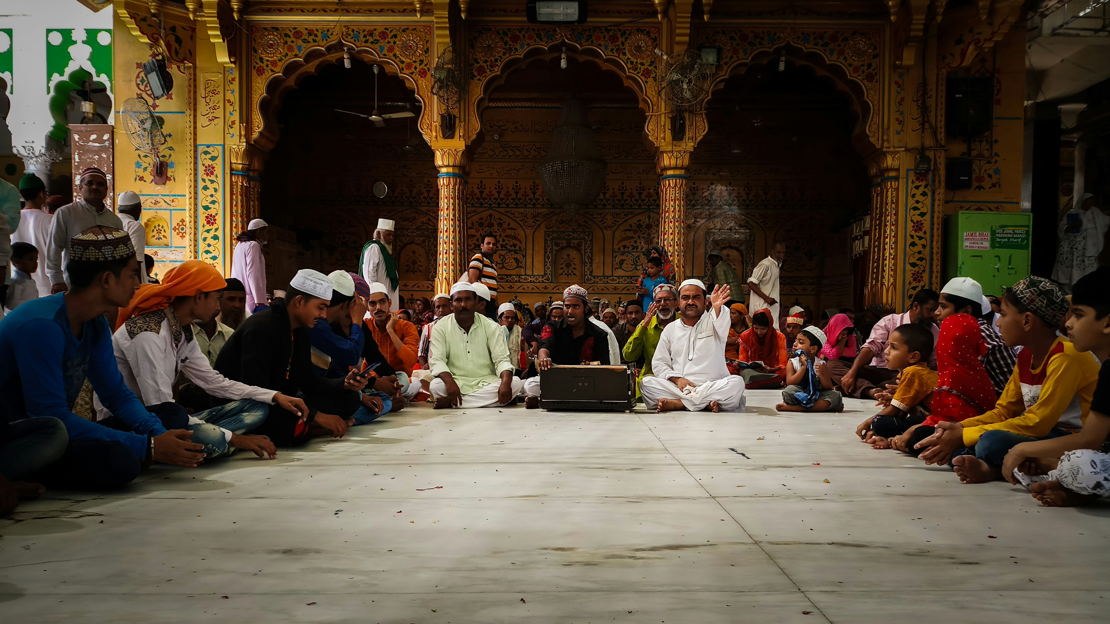 many seated people wearing robes and turbans line a central space, framed by the arches of a Mosque