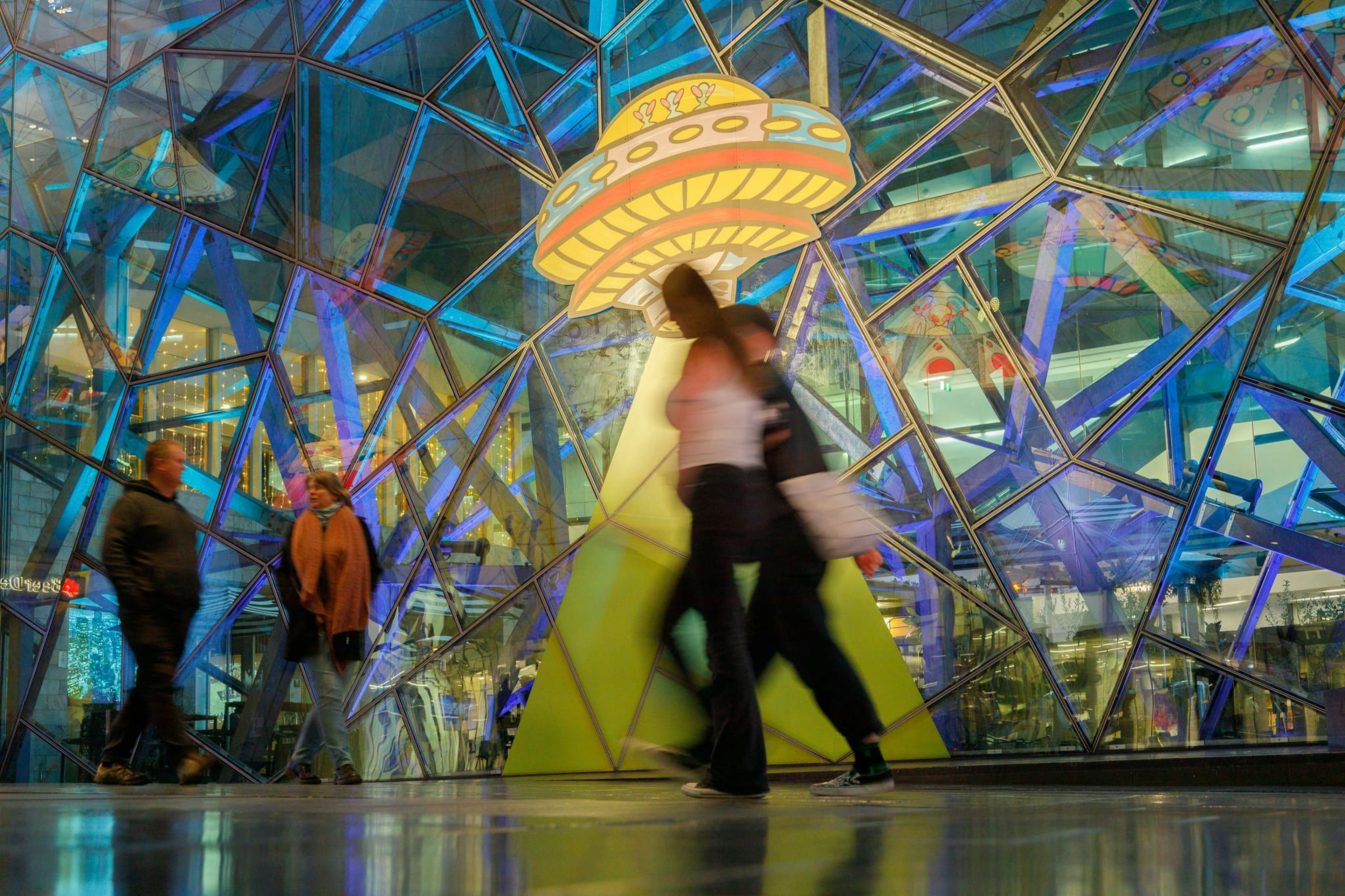 Two people walk past an image of a spaceship, with a yellow beam decaled on the glass fractal panels of The Atrium at Fed Square. They are blurry, and walk through the beam.