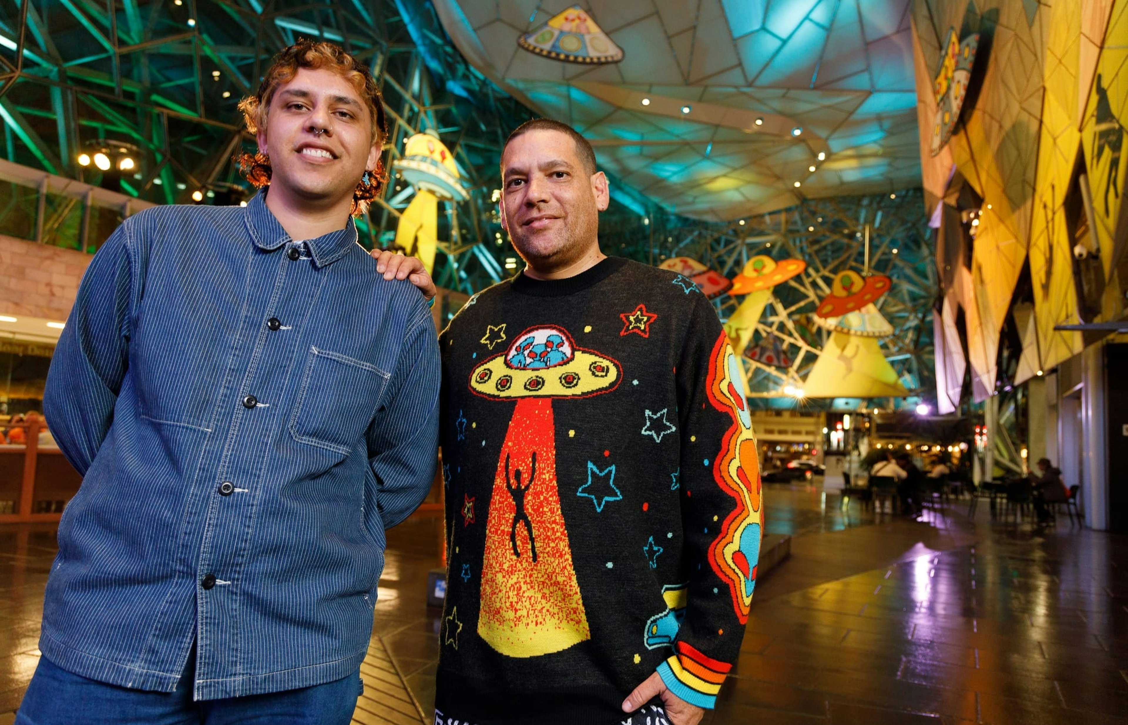 Two people look at the camera, one wears a blue shirt, the other a jumper featuring a ufo and a person being beamed up in an orange glow. Behind them is the Atrium at Fed Square, full of 2D large cut out spaceships and yellow decals on the walls showing beamed up silhouetted bodies.