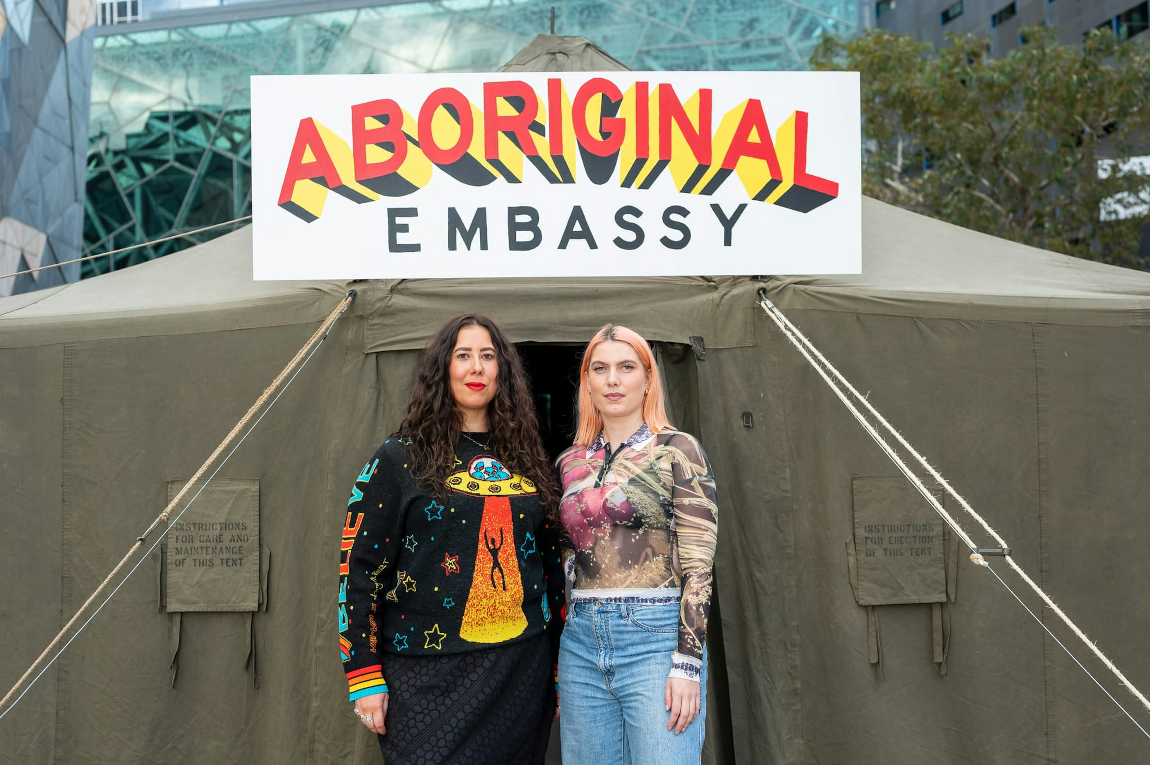 Two women stand in front of a khaki tent, the words 'Aboriginal Embassy' on a sign hangs above their heads.