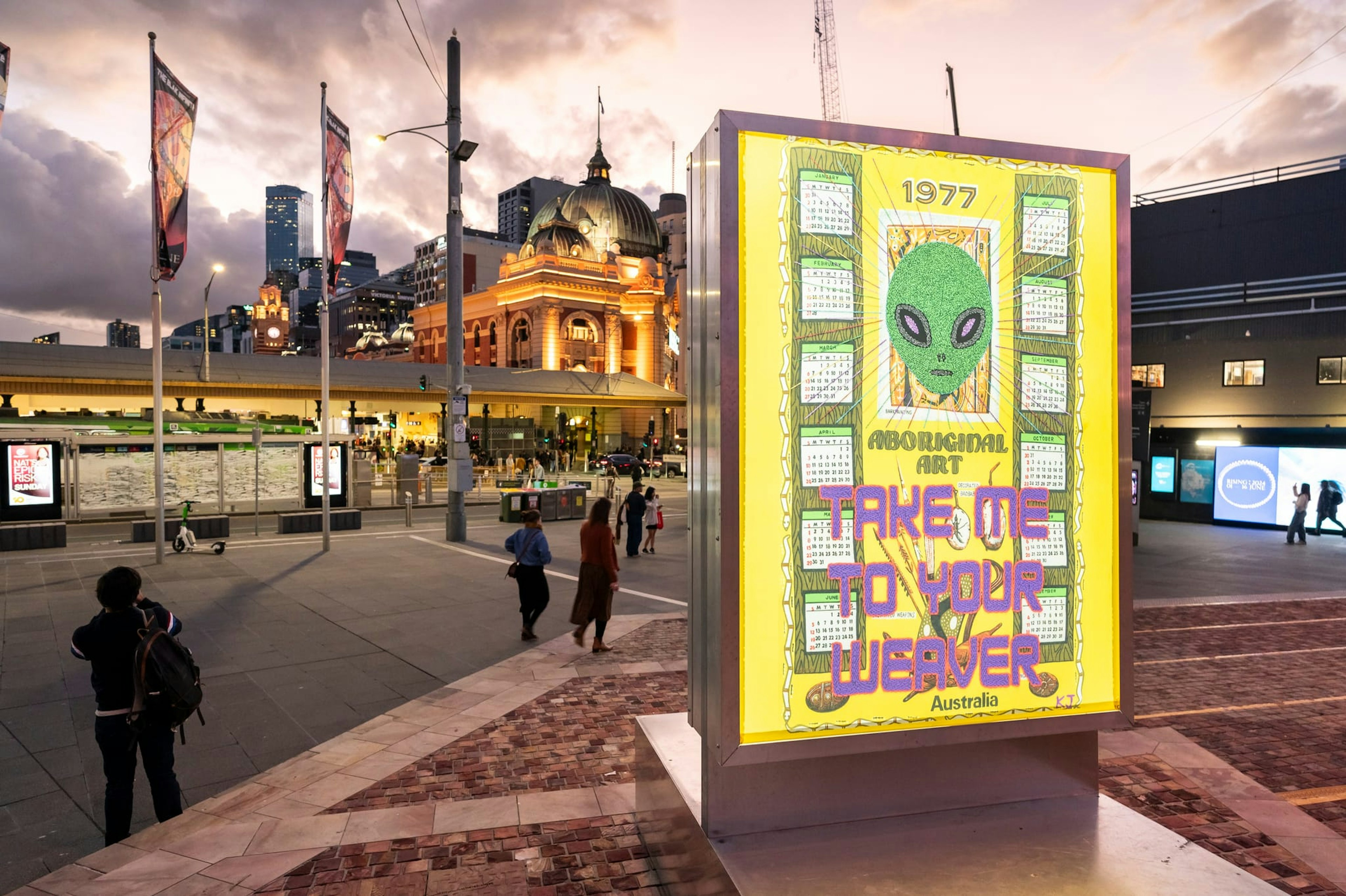 A bright yellow lightbox displays an artwork with a large green alien head and 'Take Me To Your Weaver' written in pink. The box sits on the steps of Fed Square, and Flinders Street Station is lit in yellow in the background. The sky is the dusky pink of twilight.