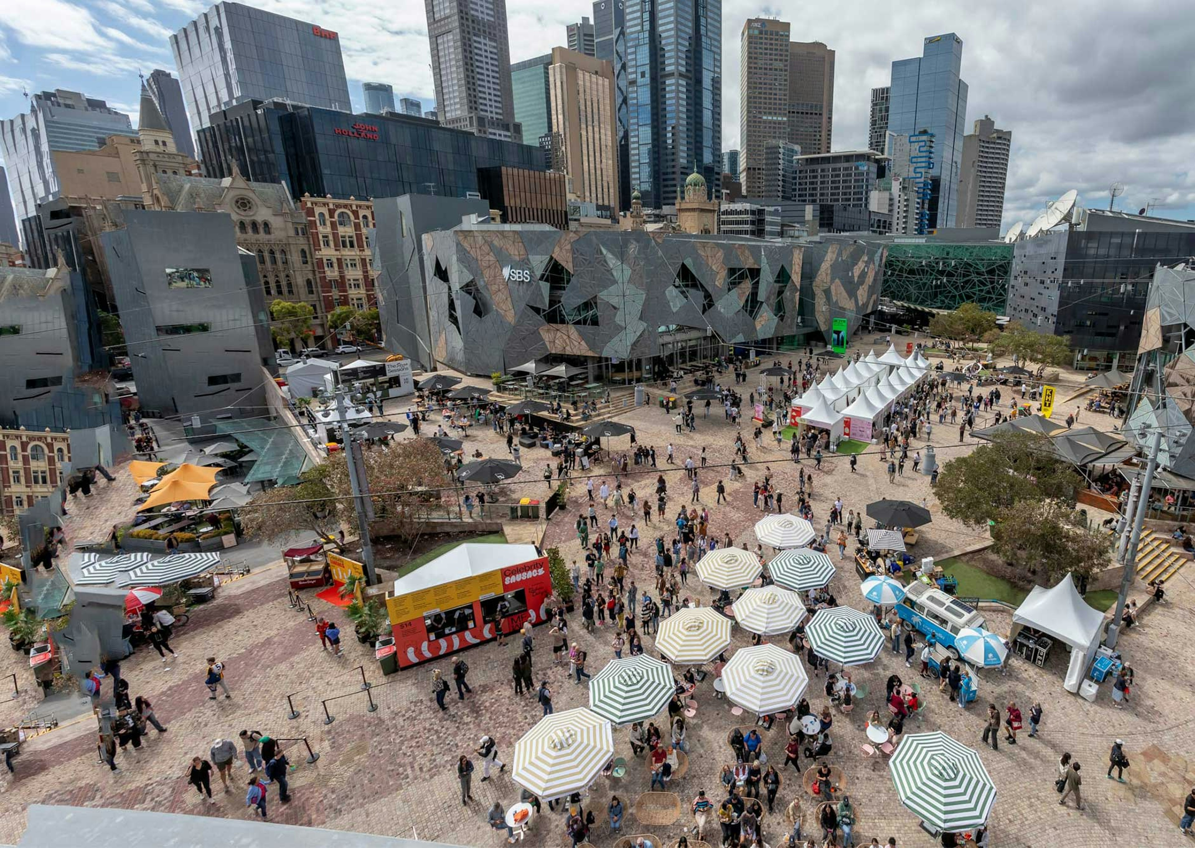 A photo from the rooftop of Transport Hotel in Fed Square, looking into the main square there is a big food festival setup with lots of umbrellas, a bar and marques with a couple of hundred people walking around