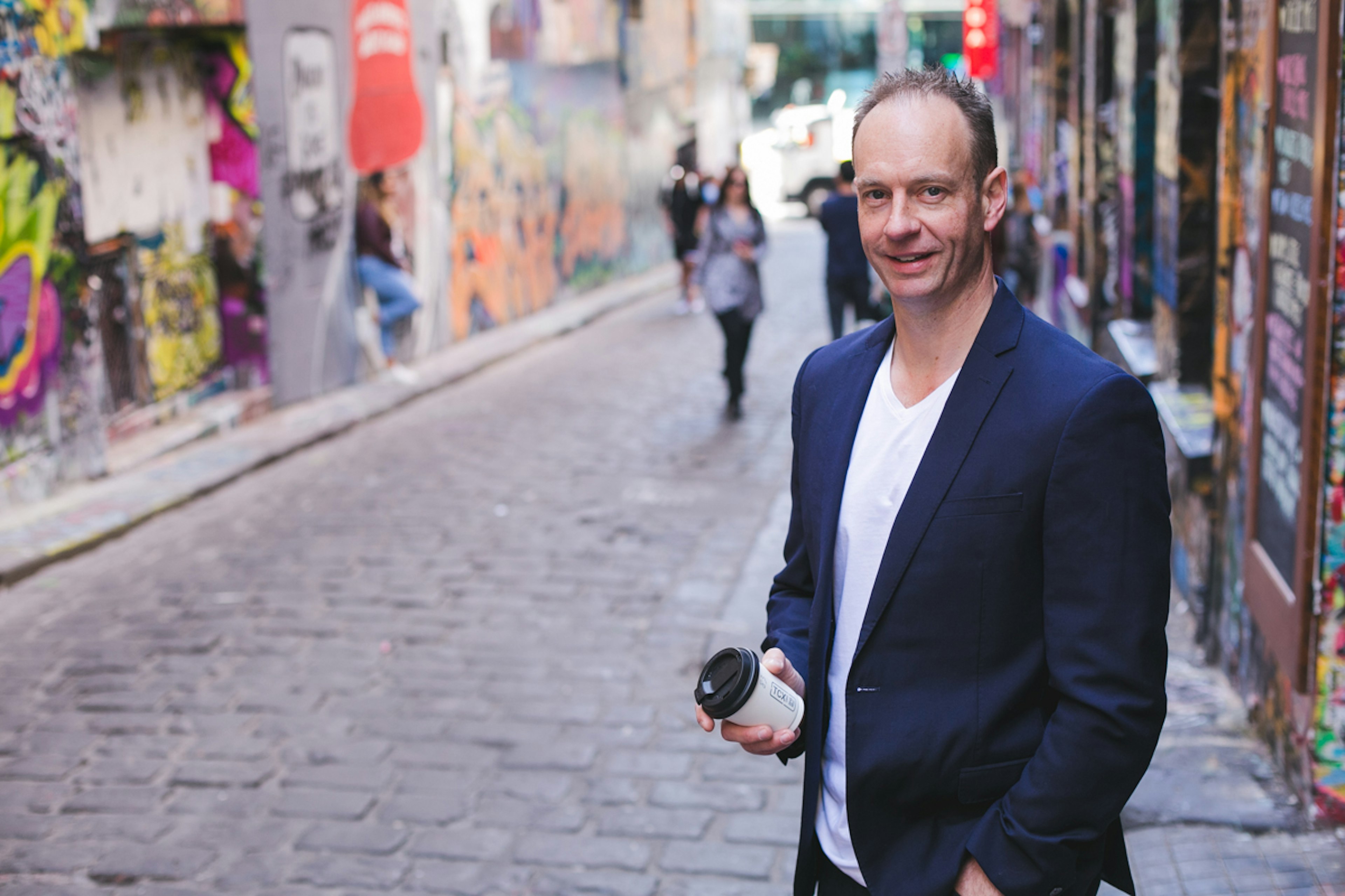 A man in a blue blazer stands in Melbourne's Hosier lane, looking at the camera and holding a coffee cup