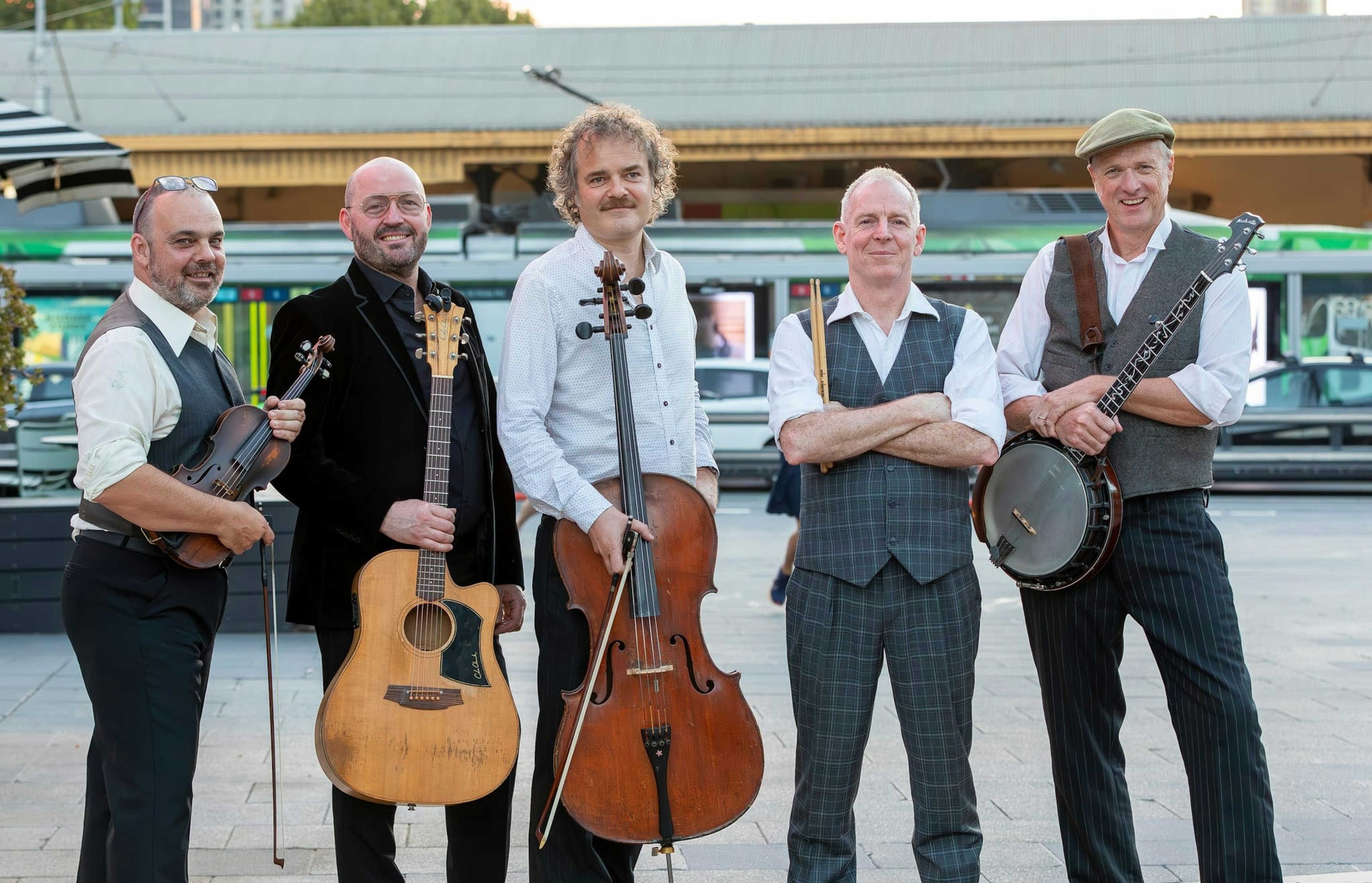 Five men dressed in great and black suits, a few with waistcoats, stand opposite Flinders Street Station, holding stringed instruments.