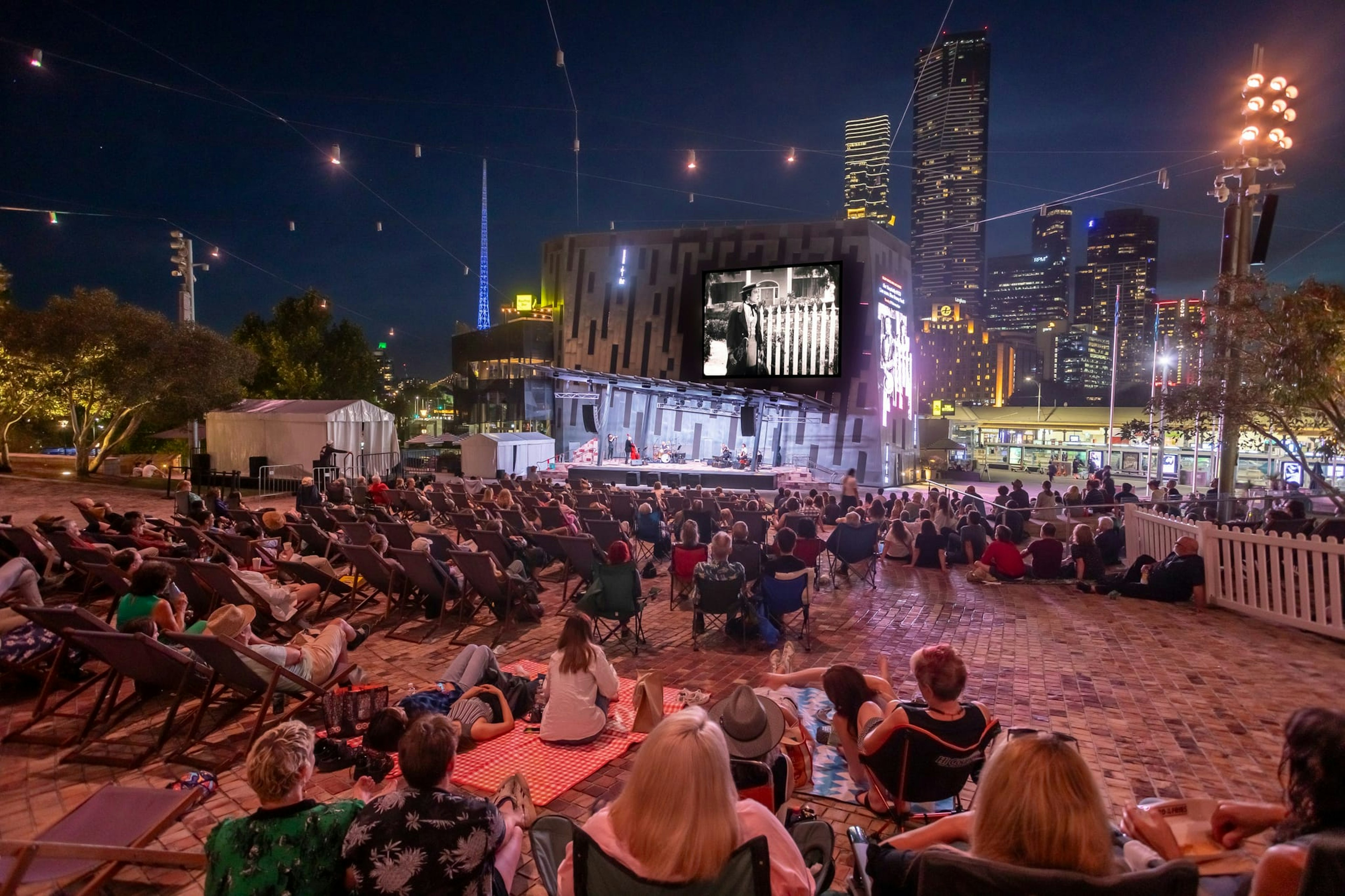 A black and white silent film plays on Fed Square's big screen at night, with hundreds of people in deckchairs watching the film. A band plays on the stage.