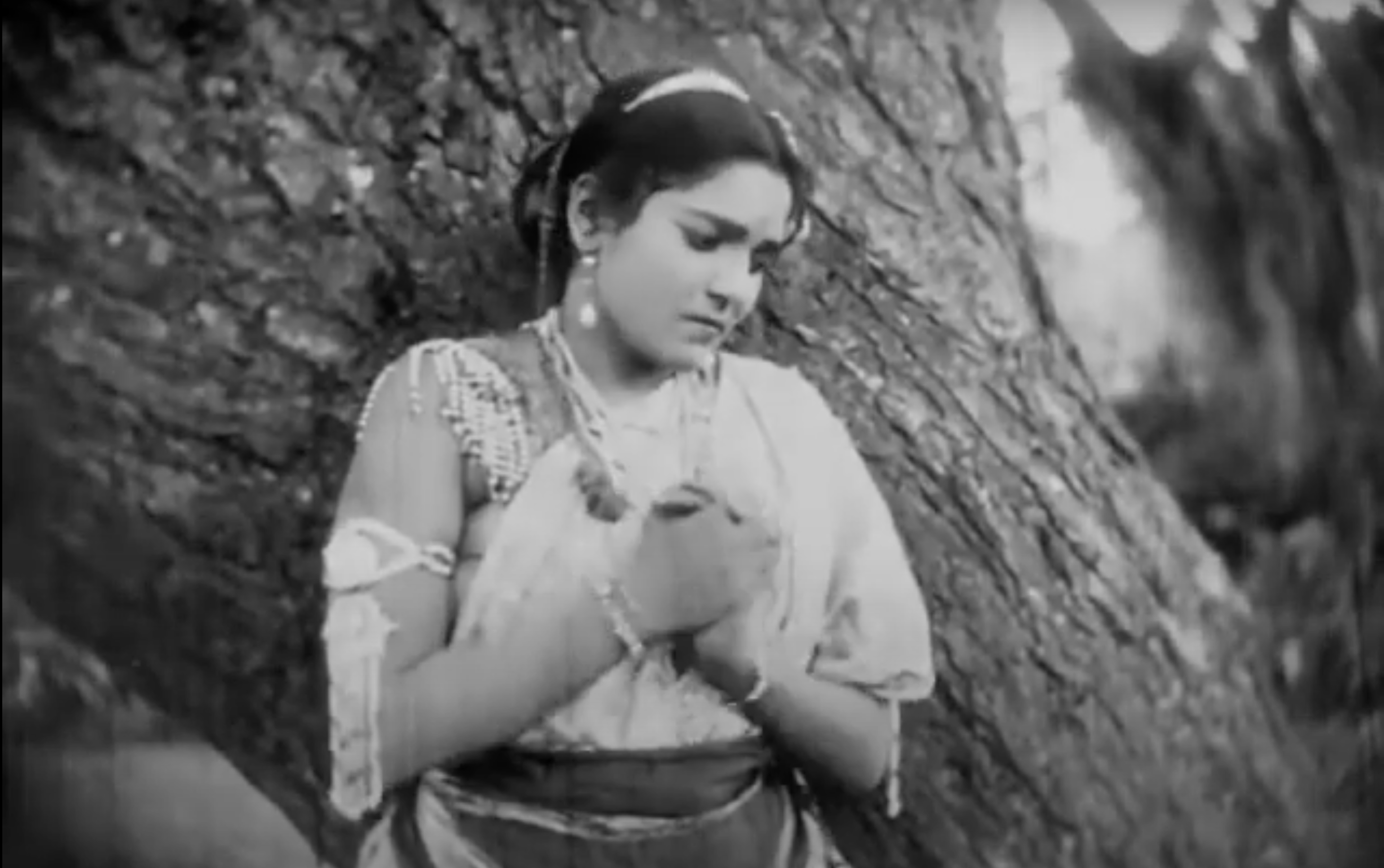 A woman in a Sari clasps her hands together, looking downward, as she leans against a tree. Black and white image.