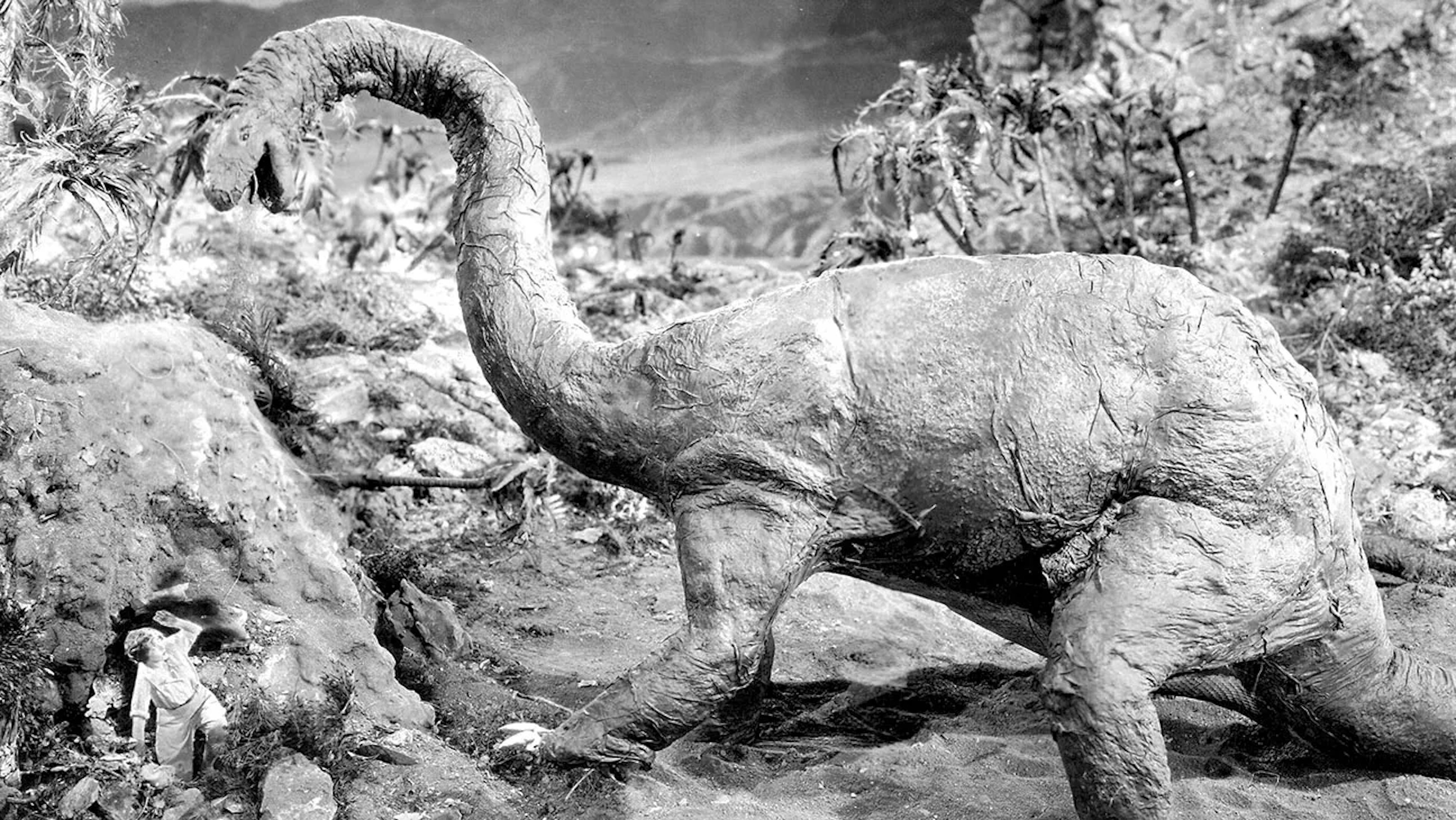 A large dinosaur with a long neck (Brontosaurus) looms over a woman, who crouches with her arm protecting her face. There are palm trees in the natural landscape around them. Black and whit image.