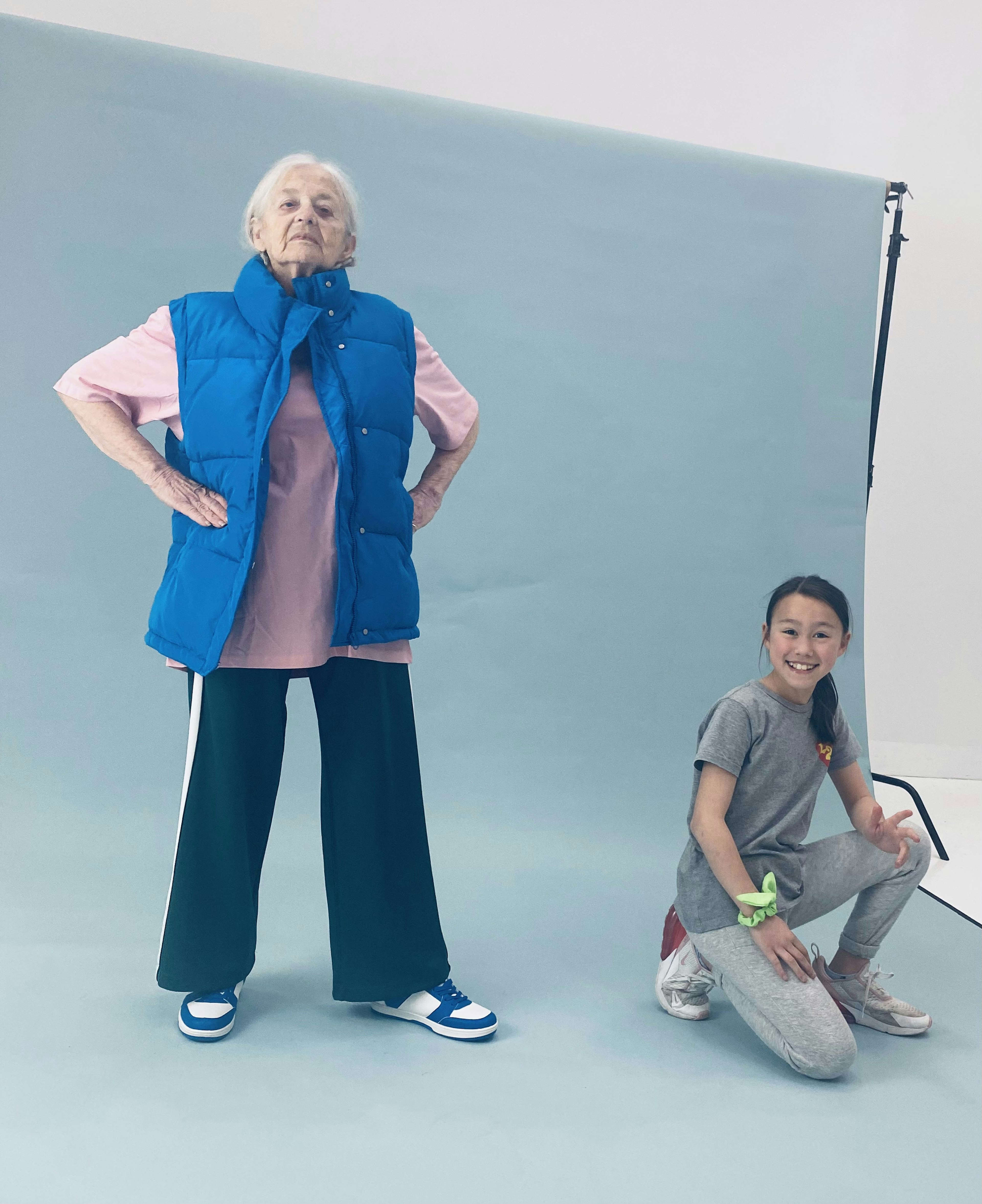 An elderly woman with a bright blue puffer jacket stands tall, with her hands on her hips, posing. Next to her, a girl in jeans kneels, posing.