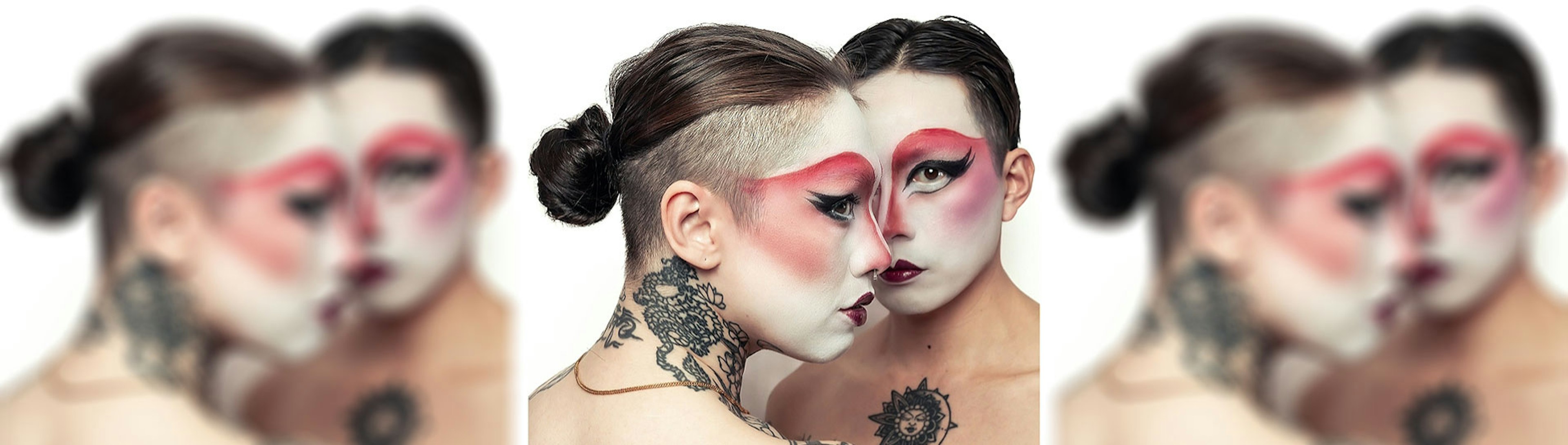 Charlie and Dyan, two genderfluid performers of Chinese descent stand face to face, their foreheads and noses almost touching. Both wear striking drag makeup inspired by Chinese opera. White base, bold red eyeshadow that sweeps across the brows, dramatic winged eyeliner, and dark lips. Their hair is slicked back, one in a low bun with shaved sides. Their bare shoulders and backs are visible, decorated with large black-and-grey tattoos — floral mandalas, an eye, a tiger with a monkey on Charlie , and a sun design on Dyan's chest. The overall effect is intense and intimate, blending softness with power.